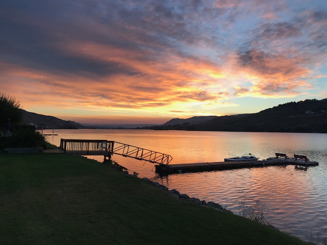 Pôr do sol sobre um lago com doca e pequenos barcos, e montanhas nos arredores.