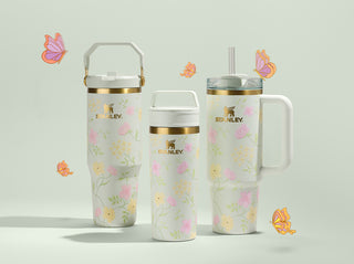 Three Stanley 1913 tumblers in white floral print with gold accents, surrounded by illustrated butterflies on a sage background.