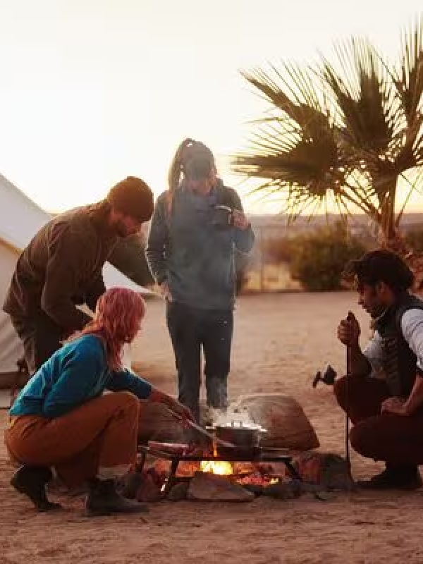 Group of friends cooking around a desert campfire at sunset near glamping tents with palm trees in background