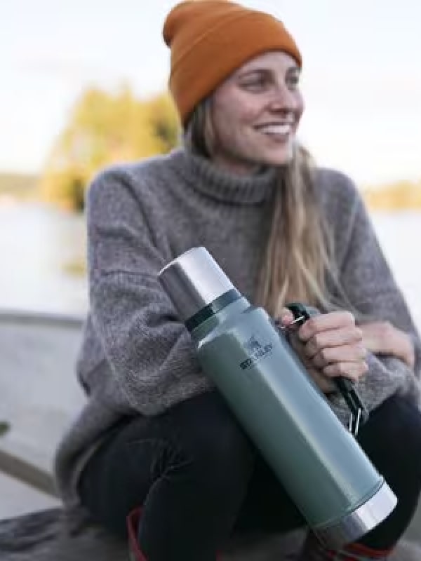 Woman smiling outdoors holding a Stanley 1913 green classic vacuum bottle, seated near a lake in autumn.