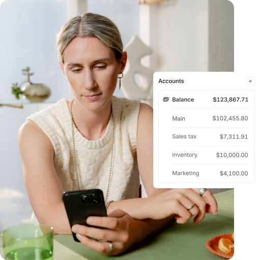 Shopify merchant checking phone with UI displaying Shopify Balance accounts for main, sales tax, inventory, and marketing.