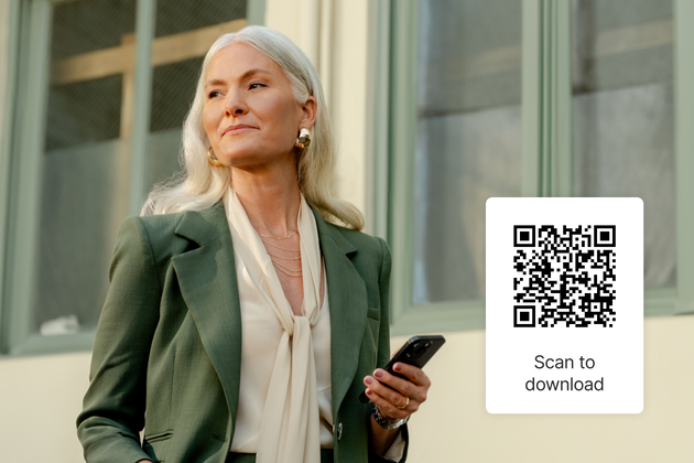 A white Shopify merchant with long silver hair wearing a stylish green blazer and a light-colored blouse stands holding a smartphone. To the left, a QR code is displayed with the text "Scan to download" beneath it.
