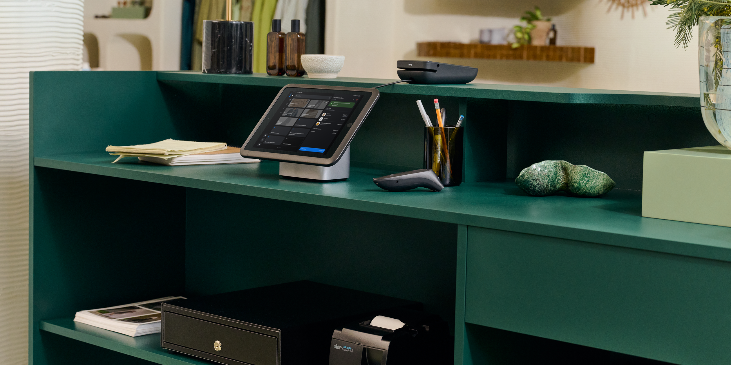 Hinter der Ladentheke ist die Verkaufsstellen-Einrichtung inklusive Tablet-Ständer, POS Terminal, Kassenschublade und Belegdrucker zu sehen.