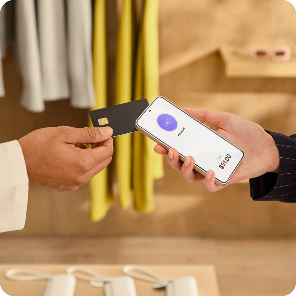 Two people are in a stylish yet warm retail environment. The customer on the left holds a credit card to tap the sales associate’s Android device to securely complete a transaction on the sales floor. The screen instructs the user to “hold card”.