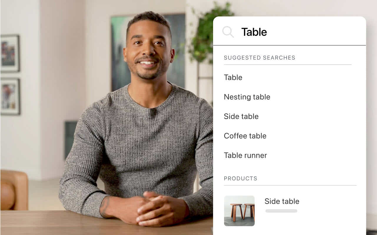 A man in a gray long sleeve shirt sits at a table and shares about the latest updates to storefront search and product recommendations. A module depicts a search bar with the word “table” in the input field. There are suggested searches for different types of tables and recommended products below the search bar.